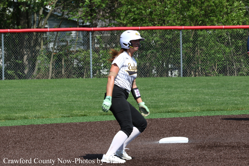 Softball player in a white helmet and striped jersey stands on a base on the infield, preparing to run during a game.