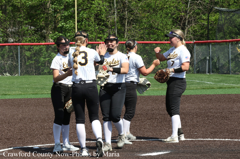 Girls softball team in white jerseys high-fiving each other on the infield dirt before a game, with green outfield and fence in the background.