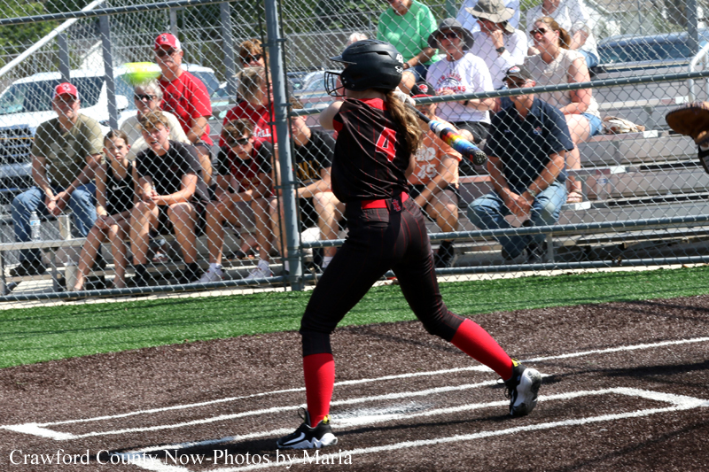 Female softball batter in black and red uniform swings at a pitch behind a chain-link fence with spectators in the stands nearby.
