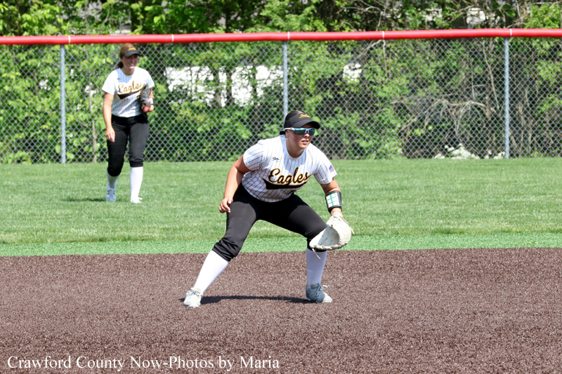 Softball infielder in a white Eagles jersey crouches to field a ground ball on the infield, with a teammate in the background.