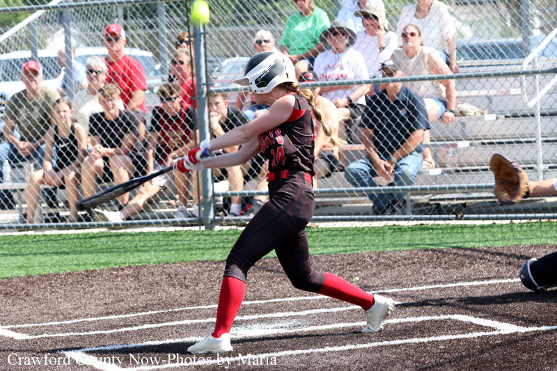 Girl softball player in a black and red uniform swings at a pitch at home plate as spectators watch from behind a chain-link fence.