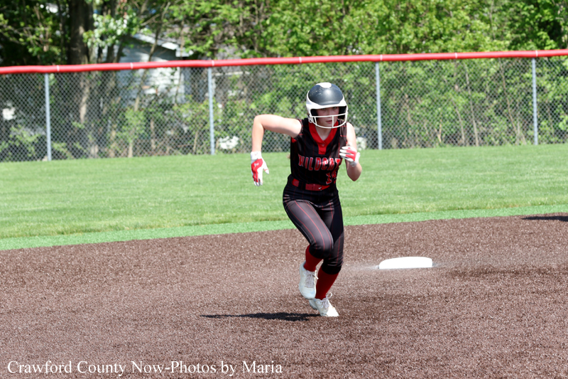 Female softball player in black and red uniform runs on the infield dirt toward a base during a game.