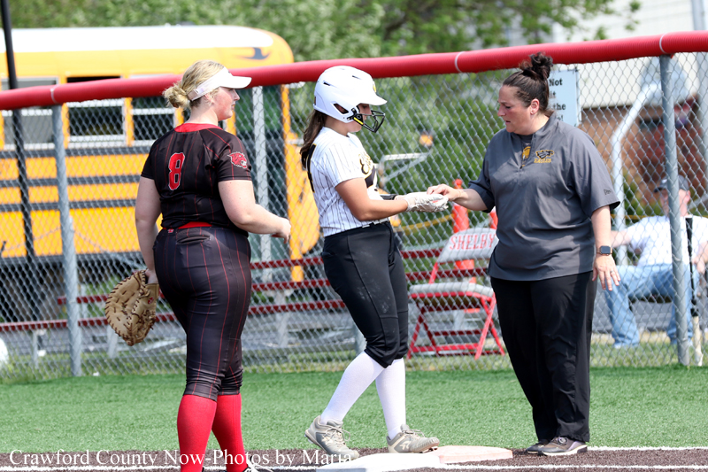 Softball base interaction: runner in white helmet receives something from an adult in gray shirt while a catcher watches nearby.