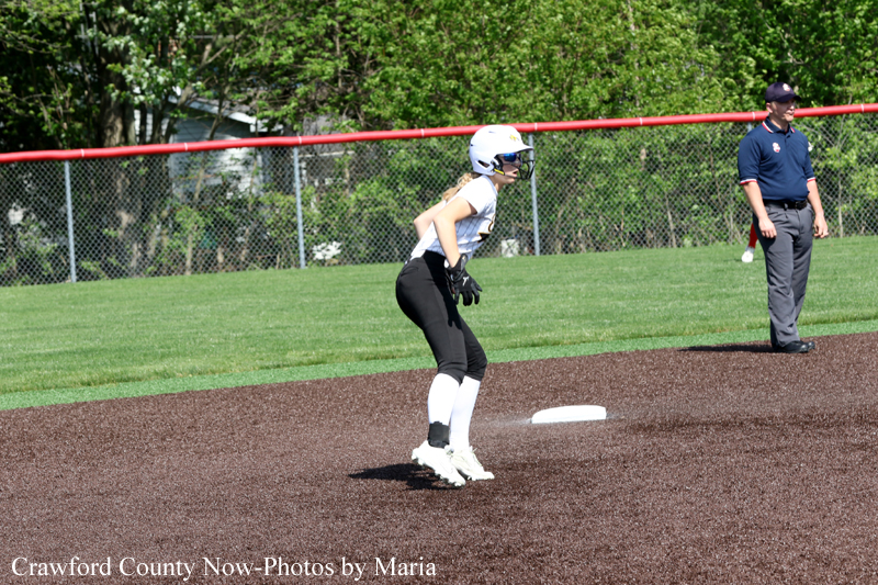 Female softball baserunner in a white helmet and black pants crouches on the dirt infield beside a base as an umpire watches from the grass in the background.