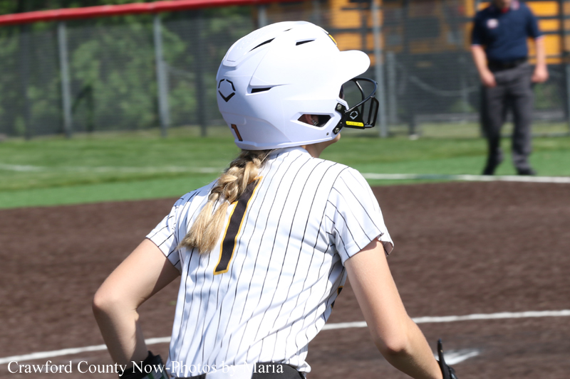 Back view of a softball player wearing a white helmet and pinstriped uniform with the number 1, standing on the infield with a braid down their back.