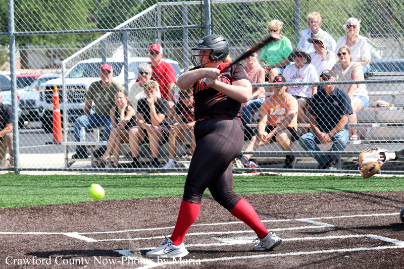 Softball batter in black and red uniform swings at a pitch on a dirt field as the yellow ball approaches; crowd sits behind a chain-link fence.