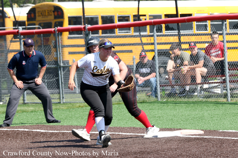 Female softball player in a white ‘Eagles’ uniform fields at first base as a opponent in red covers the bag; umpire and spectators behind the chain-link fence with school buses in the background.