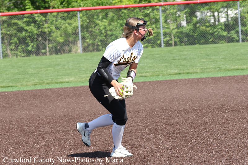 Female softball player in a white and black uniform bends to field the ball on a dirt infield, glove ready and face guard in place.