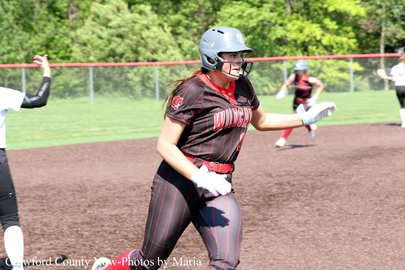 Softball player in a black pinstripe uniform and gray helmet runs across the dirt infield, white gloves on both hands.