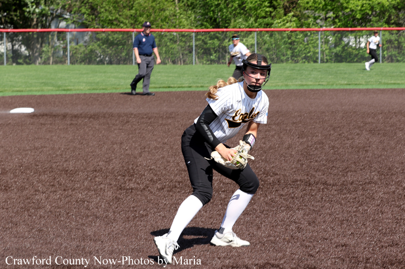 Female softball fielder in a white jersey and black pants crouches with a glove, ready for a play on the infield dirt at a game.