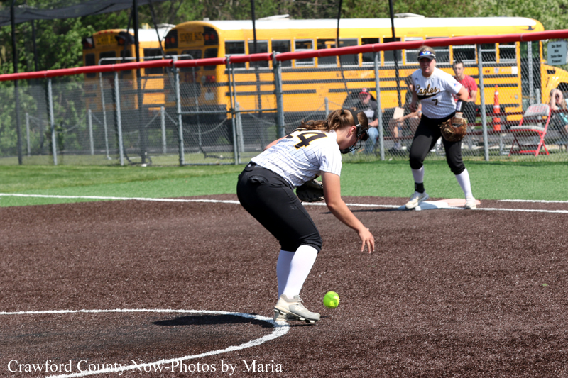 Softball player in white and black uniform bends to field a ground ball at the pitch circle; teammate near second base with yellow school buses in the background