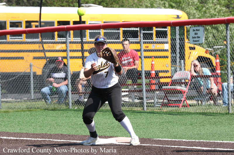 Softball player in a white jersey with number 7 prepares to catch a ball near first base on a grass-infield, with a chain-link fence and yellow school buses in the background.