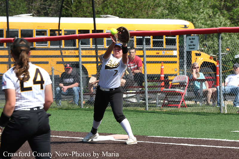 Softball player in white uniform with number 7 catching a ball with a glove at first base, near a chain-link fence and parked school buses in the background.
