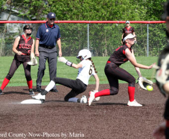 Softball game action: baserunner slides into the base as a fielder in black and red reaches for the ball, with an umpire and teammates nearby.