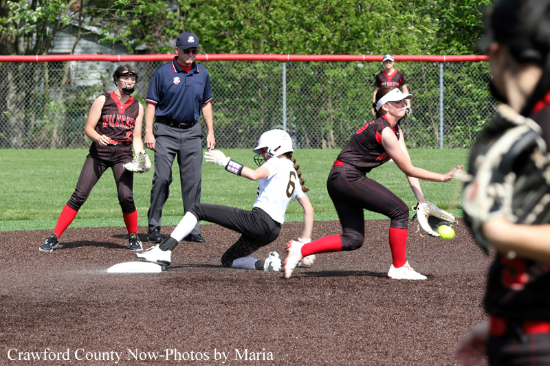 Softball game action: baserunner slides into the base as a fielder in black and red reaches for the ball, with an umpire and teammates nearby.