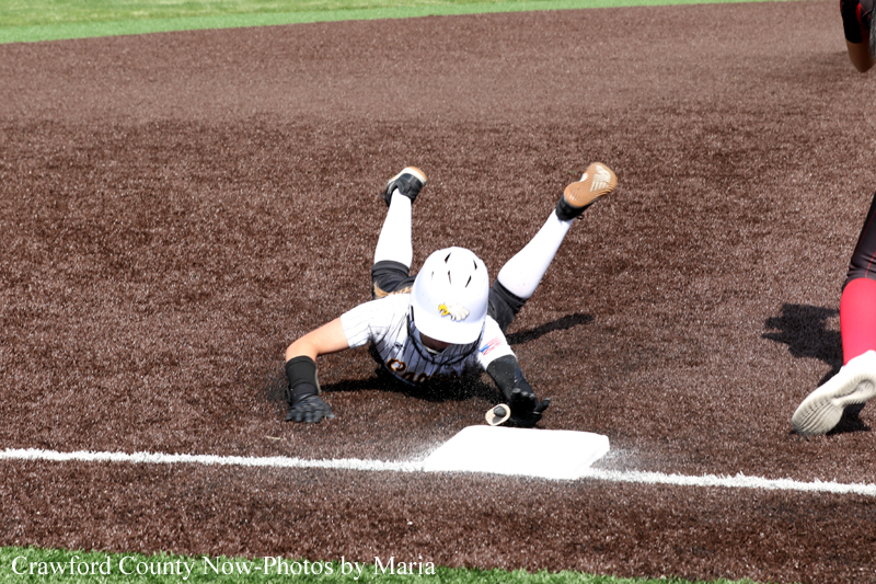 Player slides headfirst into a base, legs up in the air, reaching for the bag on a dirt infield during a softball game.