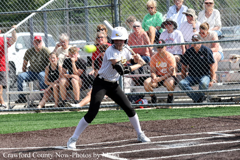 Young female softball batter in a white pinstripe uniform swings at a pitch at home plate, crowd watching from behind the fence.