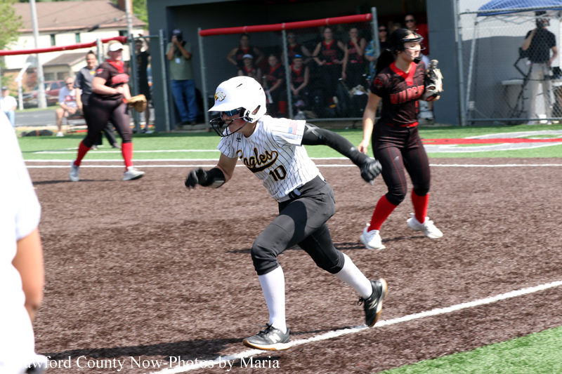 Female softball player in a white pinstripe uniform sprints along the infield, helmet on and glove ready, with a defender in black and red in the background on the field.