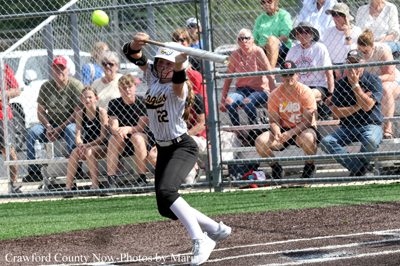 Female softball batter in a white and black uniform swings at a pitch during a game, with spectators behind a chain-link fence.