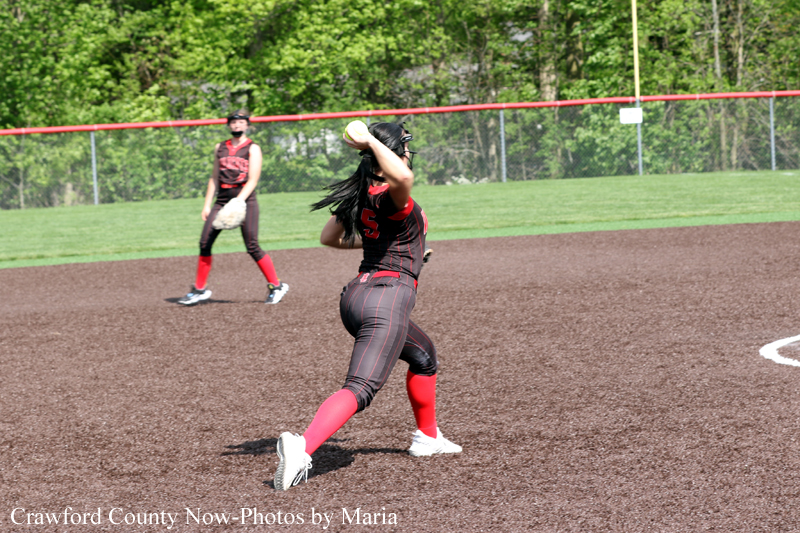 Female softball pitcher in a black-red uniform delivers a pitch on the infield, with a teammate in the background.