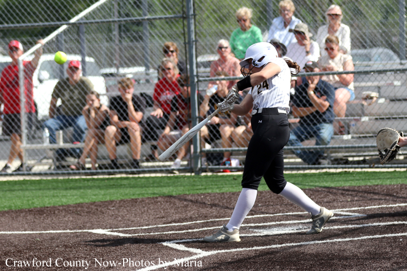 Female softball batter in white jersey and black pants swings at a pitch as the ball travels toward the plate, with spectators behind a chain-link fence watching.