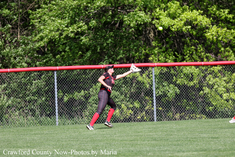 Female softball player in black and red uniform extends a gloved hand to catch a ball on a grassy outfield with a chain-link fence and green trees in the background.
