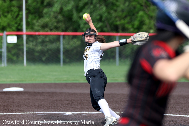 Softball pitcher in a white pinstripe jersey delivers a pitch with one leg lifted and glove extended toward the plate.