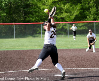 Female softball pitcher in a white pinstripe uniform winds up a pitch on the dirt mound, with a teammate and field fence in the background.