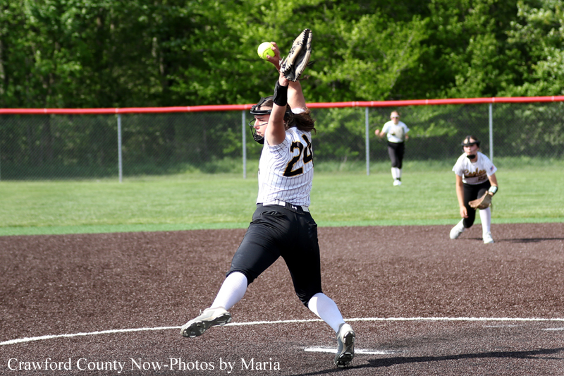 Female softball pitcher in a white pinstripe uniform winds up a pitch on the dirt mound, with a teammate and field fence in the background.