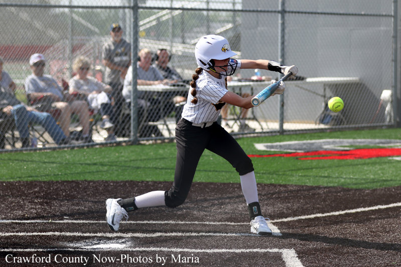 Young female softball player in a white uniform swings at a pitched ball on a fenced field, spectators watching from behind the dugout