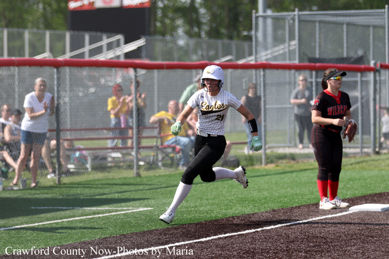 Softball player in a white Eagles uniform runs toward first base as a fielder waits at the bag, with spectators behind a chain-link fence.