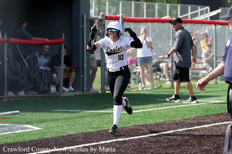 Female softball player in a white Eagles jersey number 10 runs toward home plate with arms raised as spectators watch from behind a chain‑link fence.