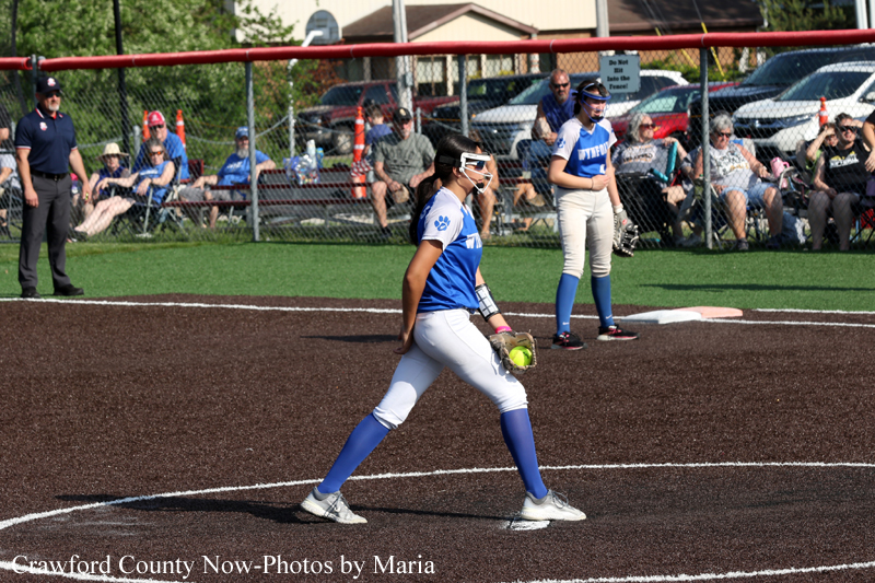 Softball pitcher in blue and white winds up on the mound, ball in glove, with spectators behind a chain-link fence in the background.