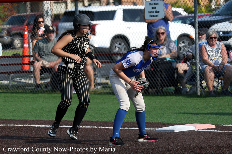 Two young softball players near first base: a runner in a black pinstriped uniform and helmet approaches as a fielder in blue and white prepares with a glove, with spectators behind the fence.