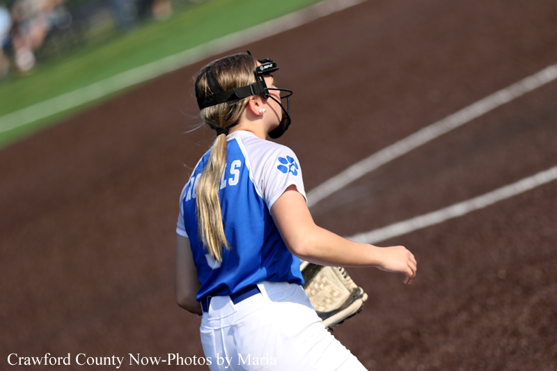 Female softball player in a blue and white uniform, facing away, wearing a protective face mask on the dirt field.