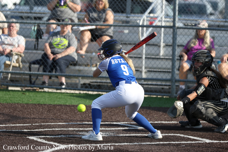 A female softball batter in a blue jersey (number 9) swings at a pitched ball while wearing a helmet and white pants, catcher nearby on dark infield dirt, fence and spectators in the stands behind.