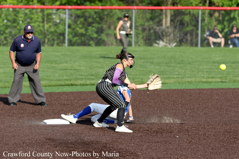 Softball catcher in pinstriped uniform and purple sleeves reaches for the ball near first base as a sliding runner and umpire look on on the infield dirt with a green outfield behind them.