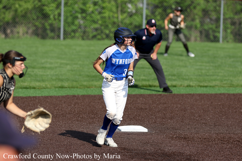 Female softball player in a blue and white uniform runs toward first base, helmet on and gloves on, as catcher and umpire watch in the background on a dirt infield.