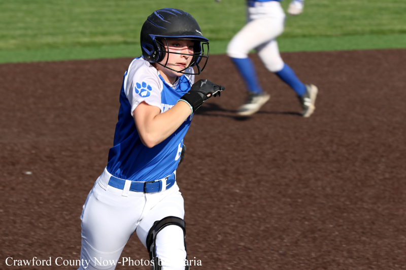 Young softball player in blue uniform runs on infield dirt, wearing a helmet and glove, focused on the play.