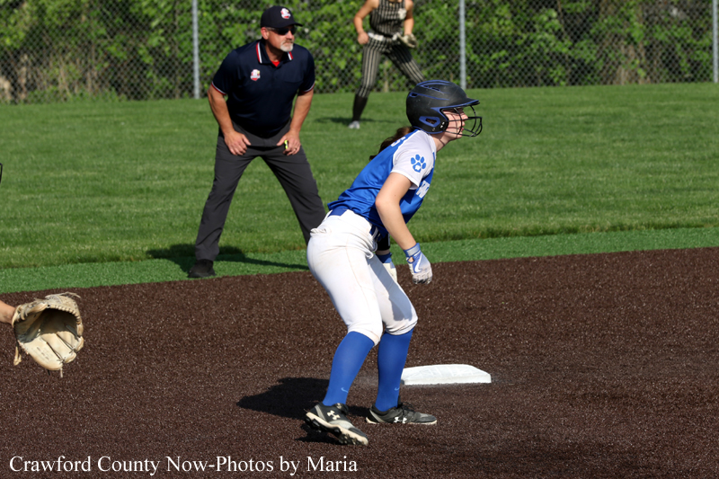 Young female softball player in blue and white uniform slides near first base while a coach watches in the background on a grassy field. Used for a play at first base scene.