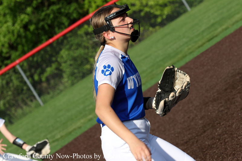 Female softball pitcher in blue and white uniform with a paw print logo, wearing a protective face mask, holding a glove on the infield dirt.
