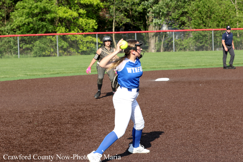 Female softball pitcher in a blue uniform winds up to throw a pitch on a dirt infield, with an umpire and green outfield in the background.