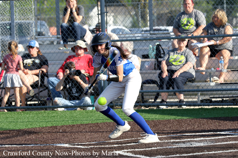 Female softball player in blue and white swings at a pitch on a dirt field with spectators behind a chain-link fence.