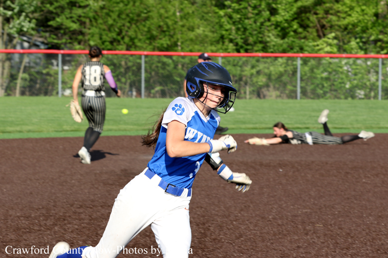 Female softball player in blue runs on the dirt infield with a helmet on, while a striped-uniform fielder stands in the background.