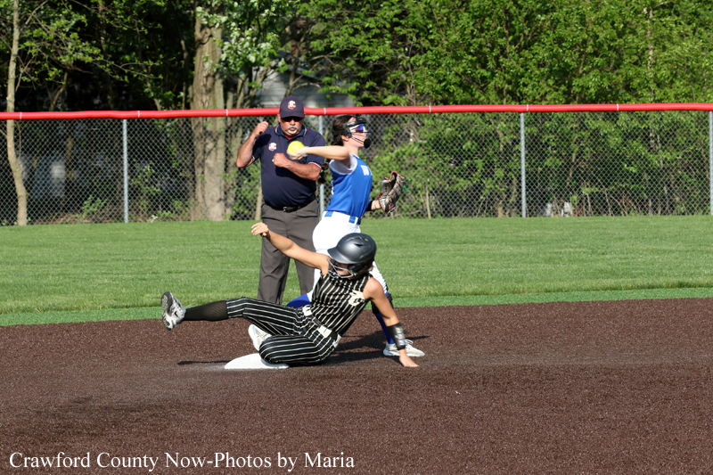 Runner slides into a base during a softball game as an opponent in blue prepares to tag; an umpire watches from behind.