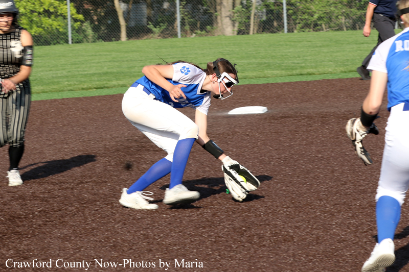Female softball player in blue and white uniform fields a ground ball with her glove near a base on a dirt infield.