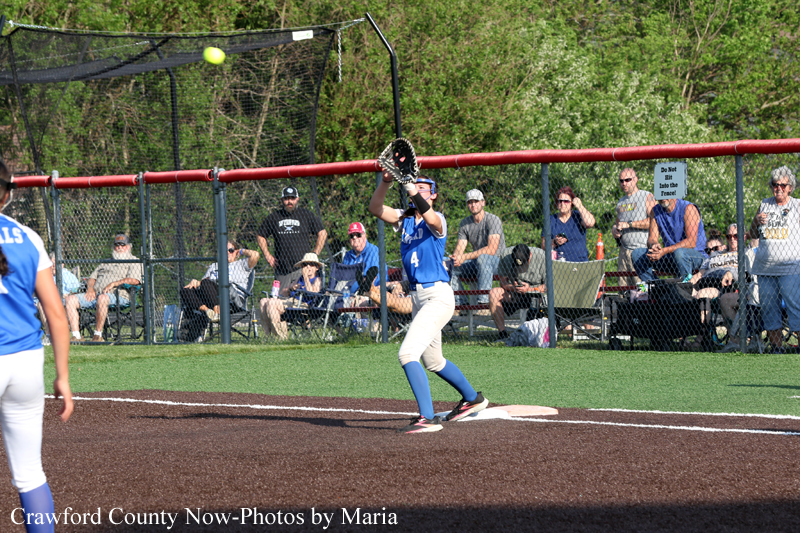 Softball batter in blue uniform at the plate, swinging as a pitch arrives; fans watch from behind the fence.