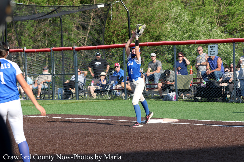 Softball pitcher in a blue uniform throwing a pitch from the mound, with spectators behind a chain-link fence watching.