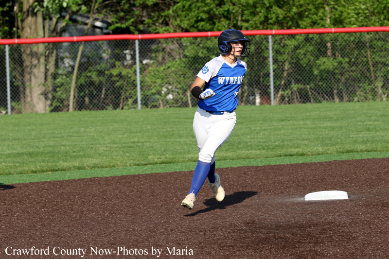 Female softball player in a blue and white uniform sprints along the dirt infield toward a base, wearing a helmet and protective sleeve with a fence and green grass in the background.