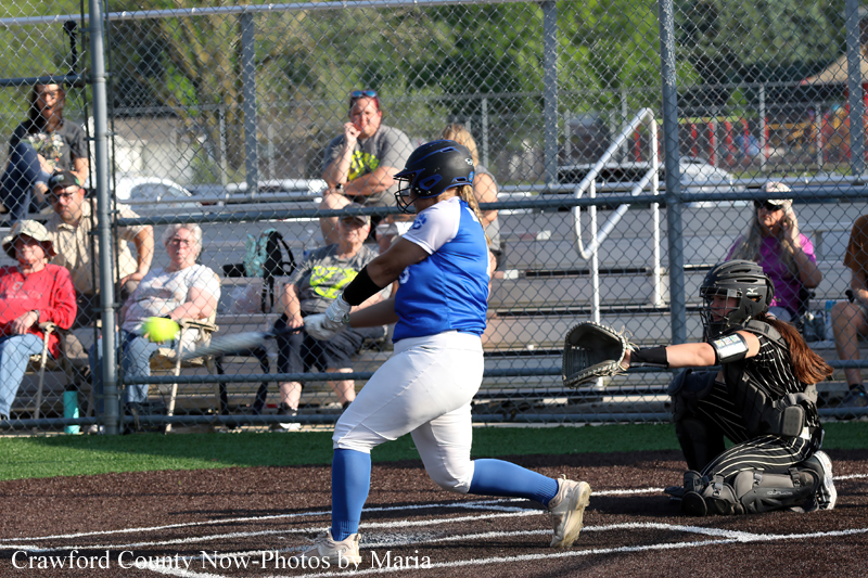 Softball batter in blue and white swings at a pitched ball as the catcher in black gear reaches for the tag; spectators sit beyond a chain-link fence.